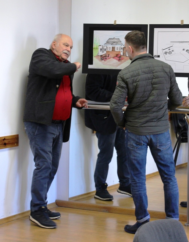 Dwóch mężczyzn stojących naprzeciw siebie obok galerii projektów wiszących na wysokości ich głów. Po lewej Krzysztof Szuster rozmawiający z Krzysztofem Małachowskim widocznym z tyłu.